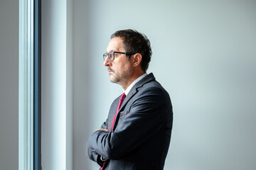 Worried businessman in elegant suit with eyeglasses looking out office window, deep in contemplation.