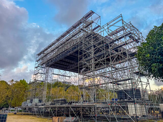 A large metal stage structure under construction in an outdoor setting. The sky is partly cloudy with trees in the background.
