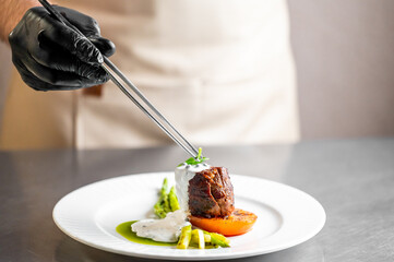 A professional chef wearing black gloves carefully garnishes a gourmet dish with tweezers in a restaurant kitchen. The fine dining plate features seared meat, asparagus, and sauce.