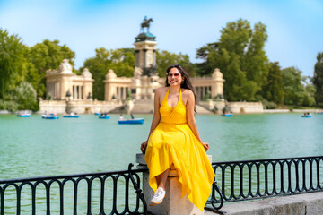 Smiling young Latina woman enjoys a relaxed moment at Retiro Park in Madrid Spain, sitting by the lake near the monument. Wears a bright yellow summer dress with white sneakers and sunglasses