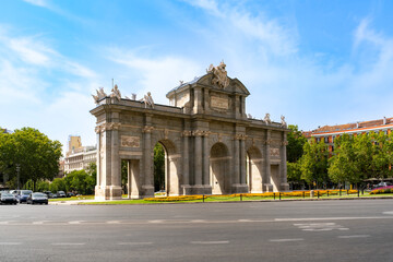 Fototapeta premium The Puerta de Alcala stands proudly in Madrid Spain under a bright summer sky. This historic neoclassical is surrounded by trees, flowers, and elegant city architecture, reflecting culture history