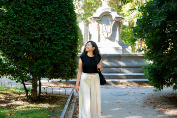 Happy young Latin woman smiling and looking up inside Museo Reina Sofía garden in Madrid Spain, wearing stylish black top and white pants with greenery and historic monument a cultural atmosphere