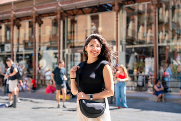 Naklejka premium Happy young Latin woman smiling outside Mercado de San Miguel in Madrid Spain, wearing casual black and white outfit, famous historic iron market a cultural icon renowned for tapas and gastronomy