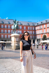 Obraz premium Happy young Latin woman smiling at Plaza Mayor in Madrid Spain, wearing stylish black top and white pants, historic red buildings and equestrian statue, representing cultural heritage and city life