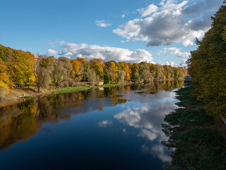 A peaceful autumn river scene with bright foliage reflecting on calm water beneath a soft blue sky.