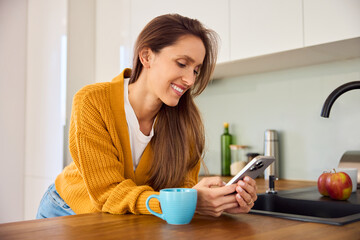 Woman in a kitchen using mobile phone while having coffee
