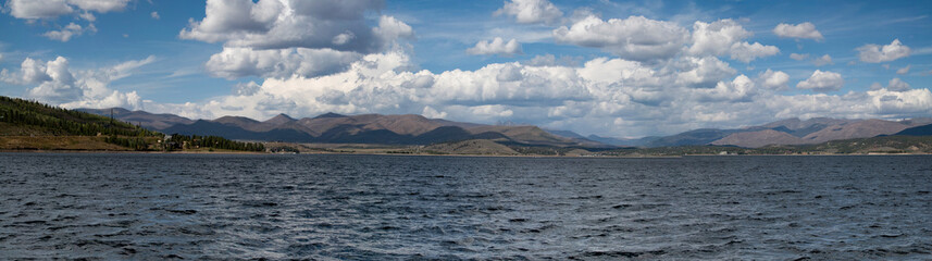 Panoramic view of the lake in Grand Lake, Colorado showing the lake in the foreground and mountains with white puffy clouds in the background.