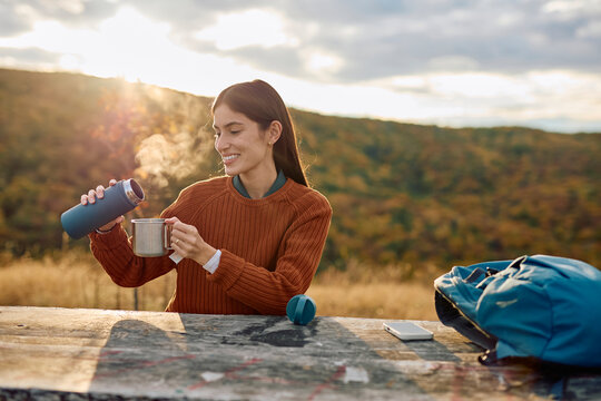 Woman pouring steaming tea from a thermos into a mug, taking a relaxing break during a hike in an autumn mountain landscape