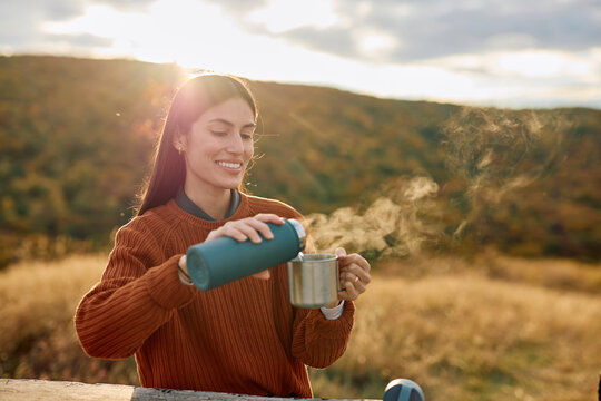 Woman standing outdoors, smiling and pouring hot beverage from a thermos into a metal cup enjoying autumn nature and warm sunlight
