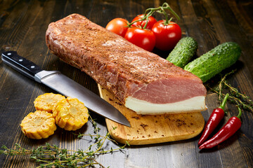 Raw seasoned pork loin with knife on wooden cutting board, surrounded by fresh vegetables and herbs. Rustic kitchen scene.