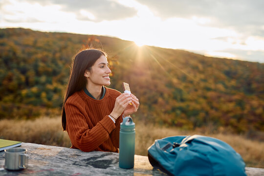 Woman taking a break from hiking, enjoying a healthy snack and smiling at the scenic view during sunset