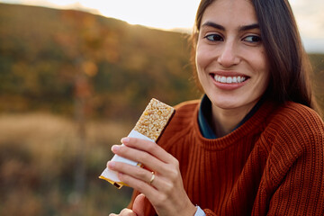 Smiling young woman eating a healthy snack bar, staying energized during an autumn hike in nature, promoting wellbeing