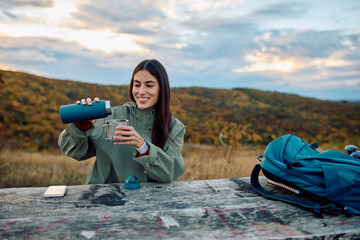 Smiling woman pouring hot beverage from thermos bottle into metal cup, enjoying a break during autumn hike in nature