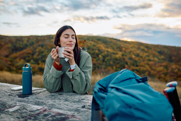 Woman relaxing and smelling hot tea from a mug while taking a break during her autumn hiking trip in nature