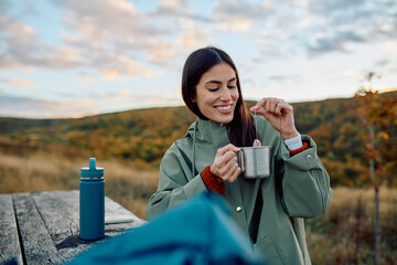 Smiling woman preparing a warm cup of tea outdoors, taking a break from hiking, enjoying autumn nature and relaxation