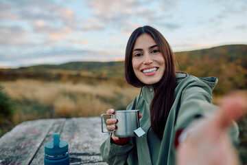 Young woman holding metal cup with tea, taking a selfie while smiling happily, enjoying nature during an outdoor activity