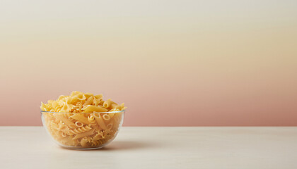 A bowl of pasta is sitting on a table. The pasta is yellow and he is dry. The bowl is clear and filled with pasta