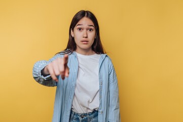 Young woman pointing at camera with surprised expression against yellow background.