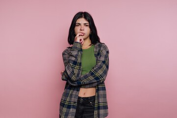 A young woman with long dark hair, wearing a plaid shirt over a green top, standing against a pink background with a thoughtful expression