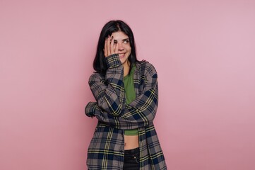Smiling woman in plaid shirt covering face on pink background.
