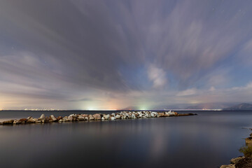 Long exposure night seascape with rocky pier and dramatic clouds over calm water