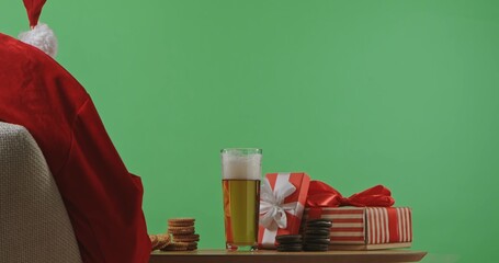 Rear view of Santa in a red suit, sitting at a table next to gift boxes, cookies, and a glass of beer. Isolated on a green background for Christmas and New Year's themes.