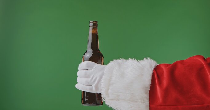 Santa Claus hand in a red suit with a white fur cuff and glove holds a dark bottle of beer, isolated on a green background. Perfect for Christmas banners, cards, and holiday advertising. - Powered by Adobe