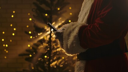 Fototapeta premium Santa Claus holding a smartphone in front of a beautifully lit Christmas tree, capturing the festive spirit. Festive mood on the eve of New Year Eve and Christmas.