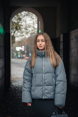 Fototapeta premium Young woman in a gray puffer jacket and black skirt walks through an urban archway on a cloudy autumn day