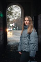 Fototapeta premium Young woman in a gray puffer jacket and black skirt walks through an urban archway on a cloudy autumn day