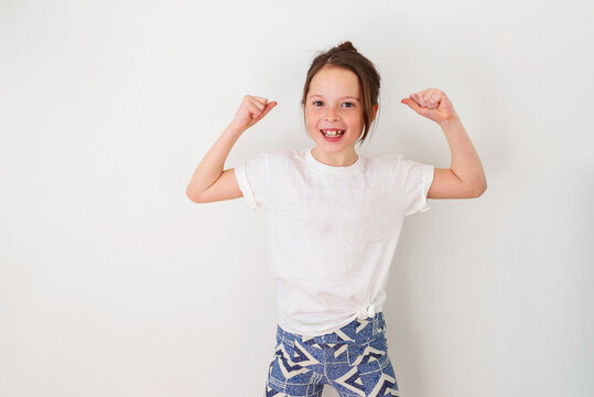 Portrait of a smiling Girl in a t-shirt standing in front of a wall flexing her muscles
