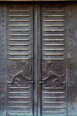 A close-up of a large, weathered metal door featuring raised slats and two sculpted lion figures facing outwards, representing the symbol of Jerusalem. The doors of the Anglo-Palestine Co. Ltd. bank, 