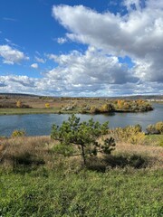 Peaceful autumn landscape with a calm river, colorful trees, and a grazing cow on a green hillside under a cloudy sky