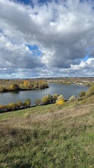Peaceful autumn landscape with a calm river, colorful trees, and a grazing cow on a green hillside under a cloudy sky