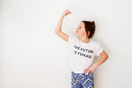 Portrait of a smiling Girl wearing a t-shirt with the slogan, the future is female standing, in front of a white wall flexing her muscles