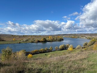 Peaceful autumn landscape with a calm river, colorful trees, and a grazing cow on a green hillside under a cloudy sky