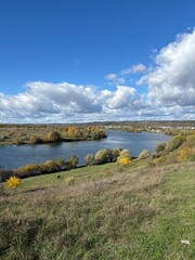 Peaceful autumn landscape with a calm river, colorful trees, and a grazing cow on a green hillside under a cloudy sky