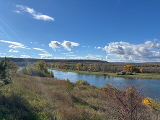 Peaceful autumn landscape with a calm river, colorful trees, and a grazing cow on a green hillside under a cloudy sky