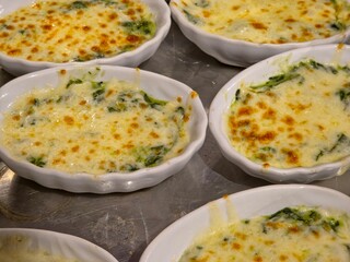 Close-up, high-angle shot of several individual white oval ceramic dishes containing a baked appetizer, likely Spinach lasagna, topped with a bubbling, golden-brown layer of melted cheese.