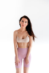 Obraz premium Sports woman standing in activewear with abs muscles, looking at camera isolated on white background. Caucasian 30s fitness girl wears beige top, pink leggings. Slim fit smiling brunette, blue eyes.
