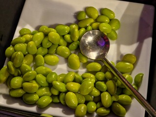 Close-up, top-down view of a pile of bright green edamame beans (shelled soybeans) on a white square platter, with a metal serving spoon resting in the lower right portion of the beans.