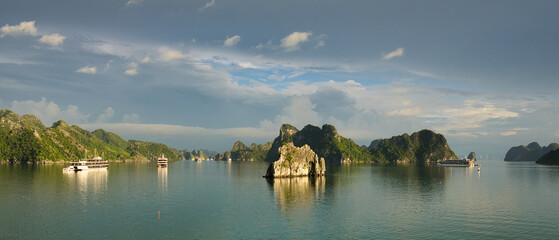 Obraz premium Ha long bay limestone karsts rise from calm emerald waters as traditional cruise ships glide through a serene, partly cloudy seascape iconic vietnam unesco scenery
