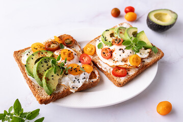 Tasty homemade toasts for breakfast with fried eggs, cream cheese, avocado, cherry tomatoes and fresh oregano leaves on white marble background top view.
