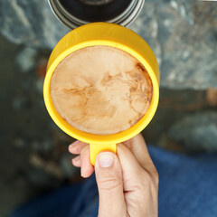 Top view of hand holding yellow mug with swirling coffee against stone background.