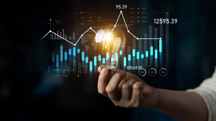 A hand holds a glowing light bulb against an overlay of business graphs, representing innovative...