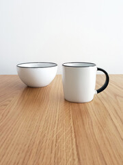 White mug and bowl set on wood and a white wall background. Front view of elegant, minimalist bowls. Ceramic household utensils. Close-up of a white stoneware bowl and mug.