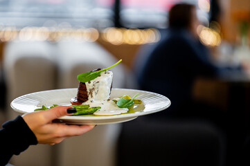 A gourmet dish of beef with a creamy white sauce and green vegetables is held on a white plate by a server in a restaurant setting.