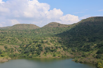 A stunning panoramic landscape of a serene lake nestled amongst rolling green hills under a bright blue sky with white cumulus clouds.