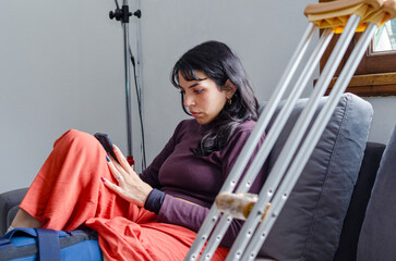 Disabled woman with a broken leg using her phone sitting on her couch next to her crutch.