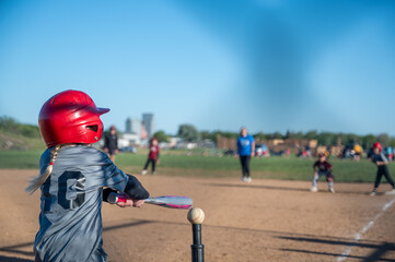 Young girl in red helmet prepares to hit baseball during practice on sunny day in park near city